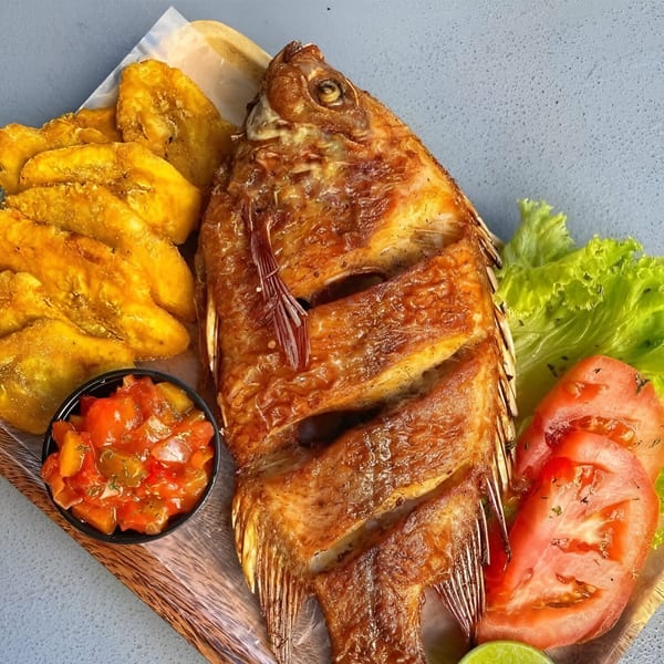 Fried whole fish with plantains, tomato, lettuce, and a side of salsa on a wooden board.