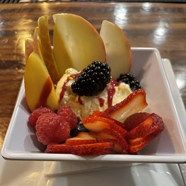 A white bowl filled with vanilla ice cream, fresh berries, and sliced apples, drizzled with sauce.