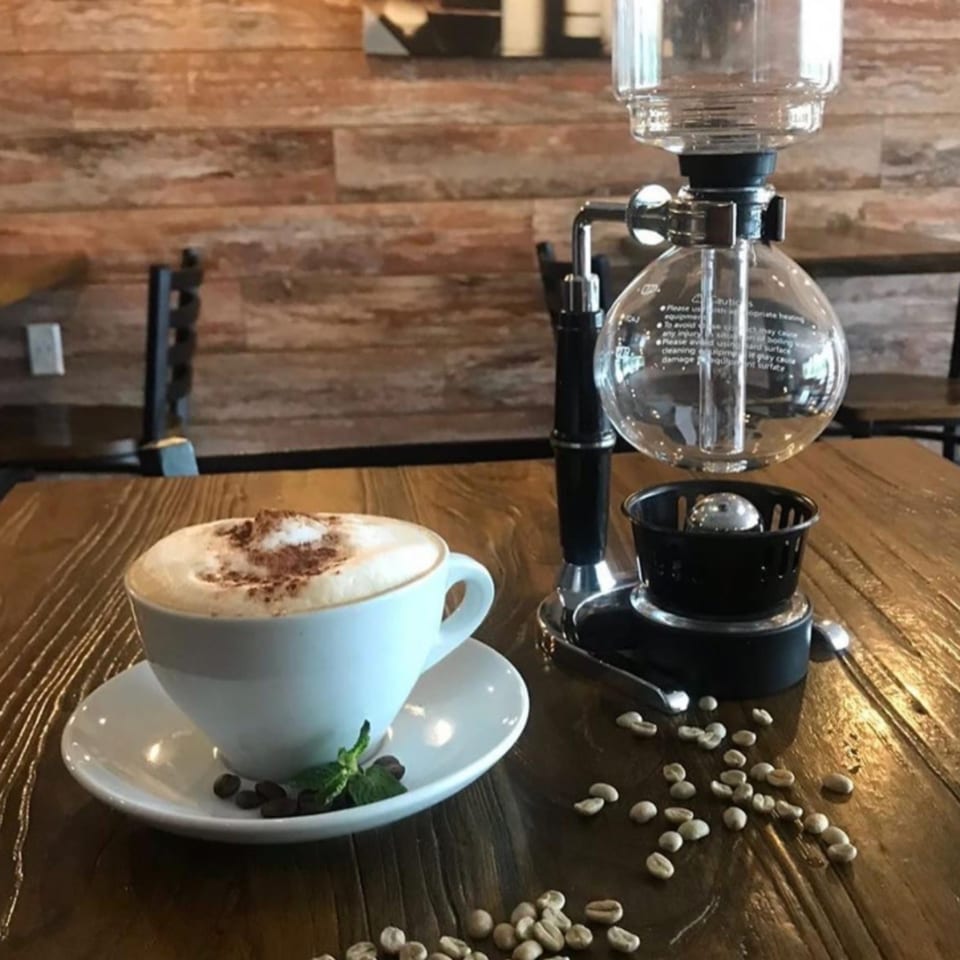 A cappuccino with a siphon coffee maker and scattered coffee beans on a wooden table.