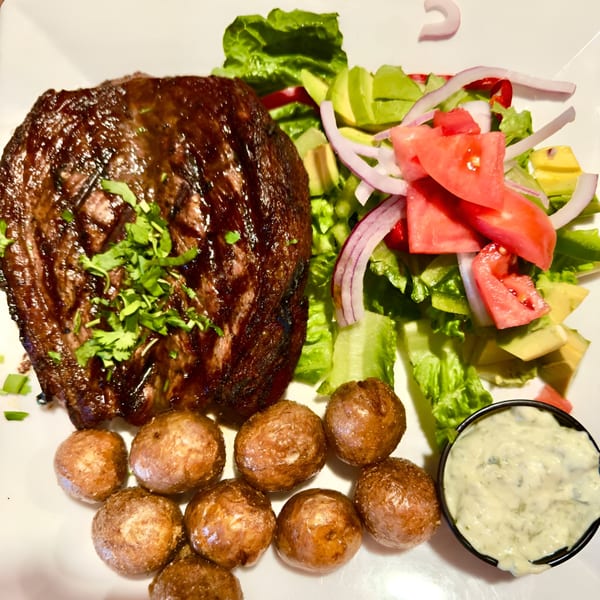Colombian steak with cilantro, roasted potatoes, a fresh salad, and a side of creamy sauce.