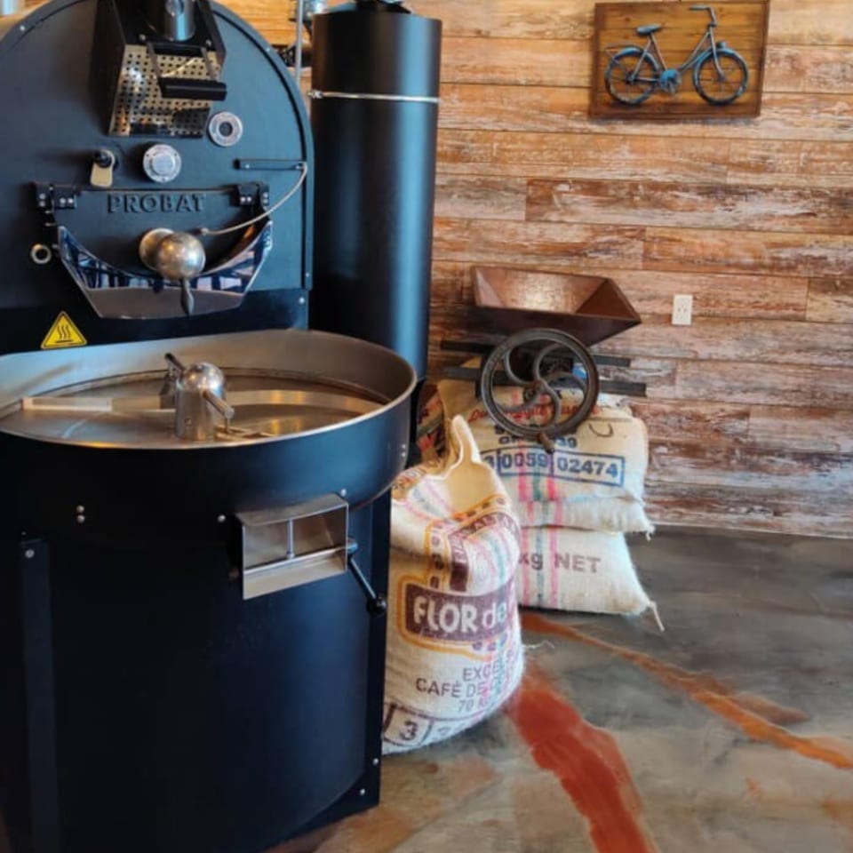 A large black coffee roaster next to sacks of Colombian coffee beans and a wooden wall.